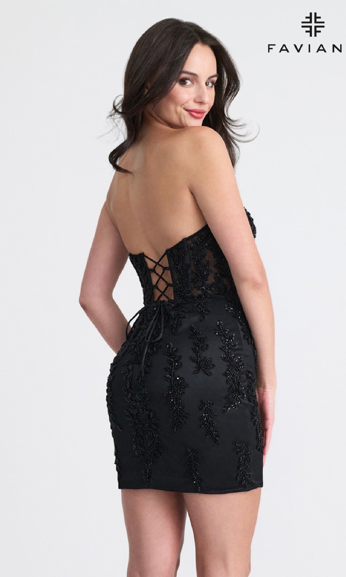 A woman in the Faviana 11317 Strapless Short Corset Hoco Dress, featuring black lace and a lace-up corset back, stands looking over her shoulder. Perfect for special occasions. The FAVIAN logo is shown in the top right corner.